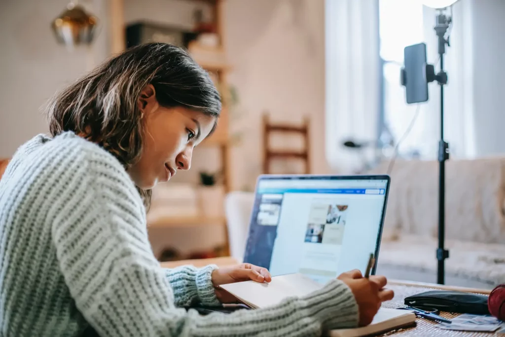 A hispanic woman is sitting at a laptop and taking notes on her laptop as she identifies Spanish business terms that will help her business.